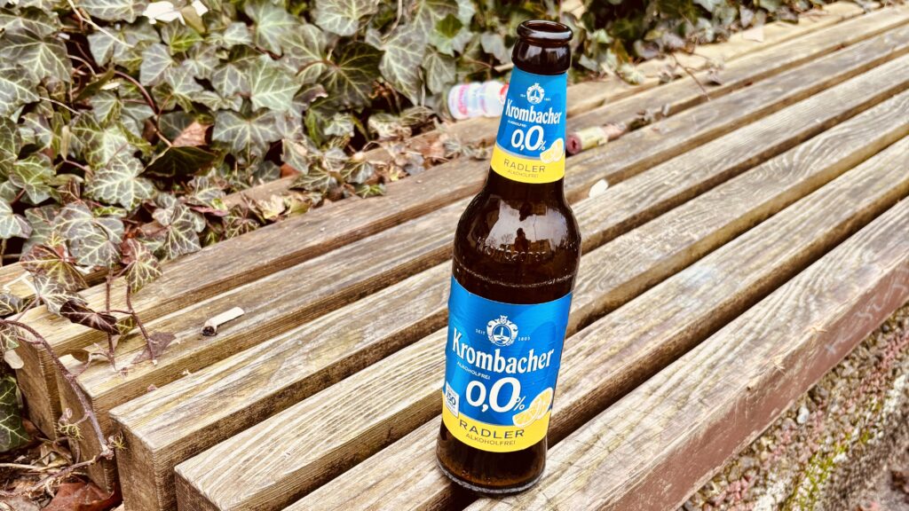 Alles falsch - Eine Krombacher 0,0% Radler Bierflasche steht auf einer Holzbank, mit Efeu und Laub im Hintergrund.