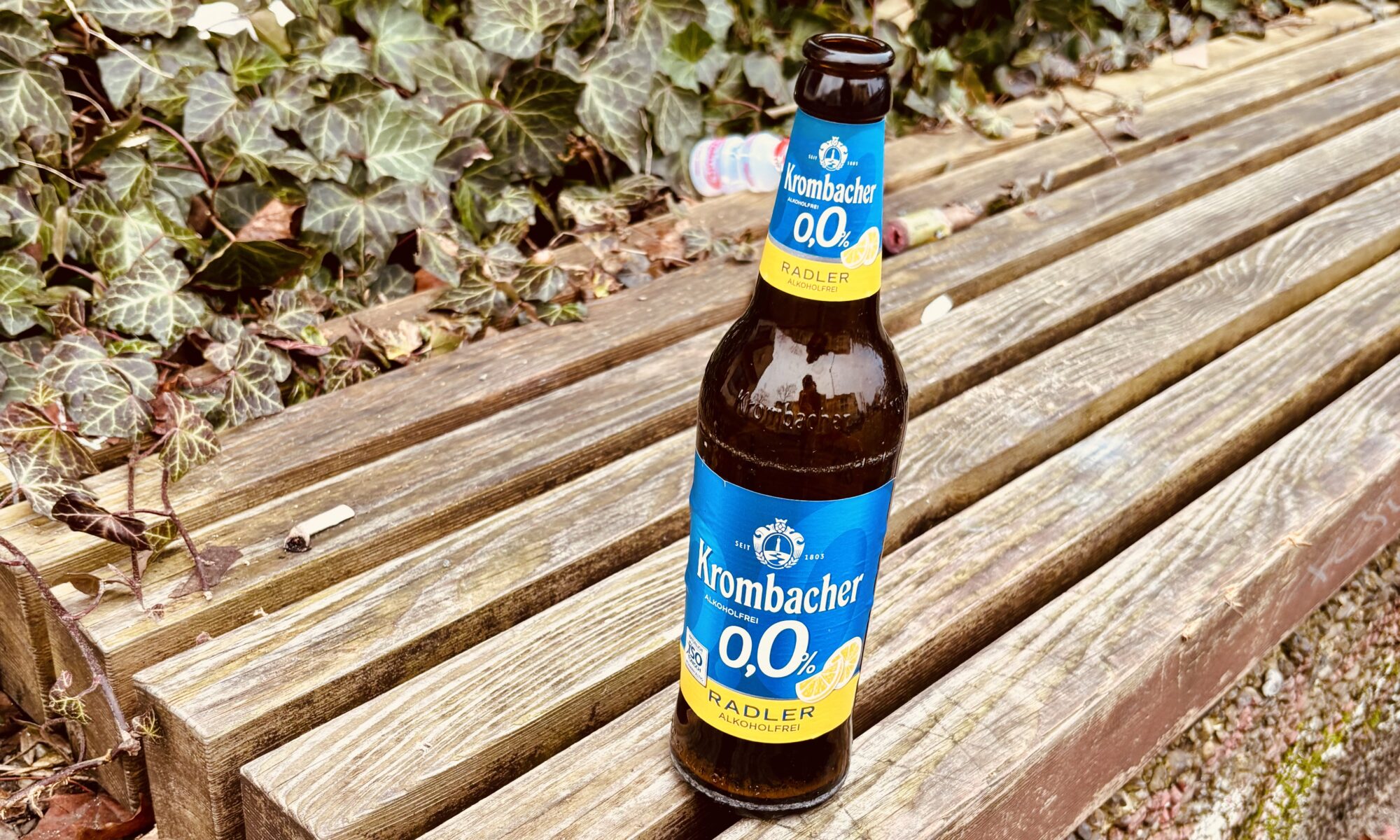 Alles falsch - Eine Krombacher 0,0% Radler Bierflasche steht auf einer Holzbank, mit Efeu und Laub im Hintergrund.