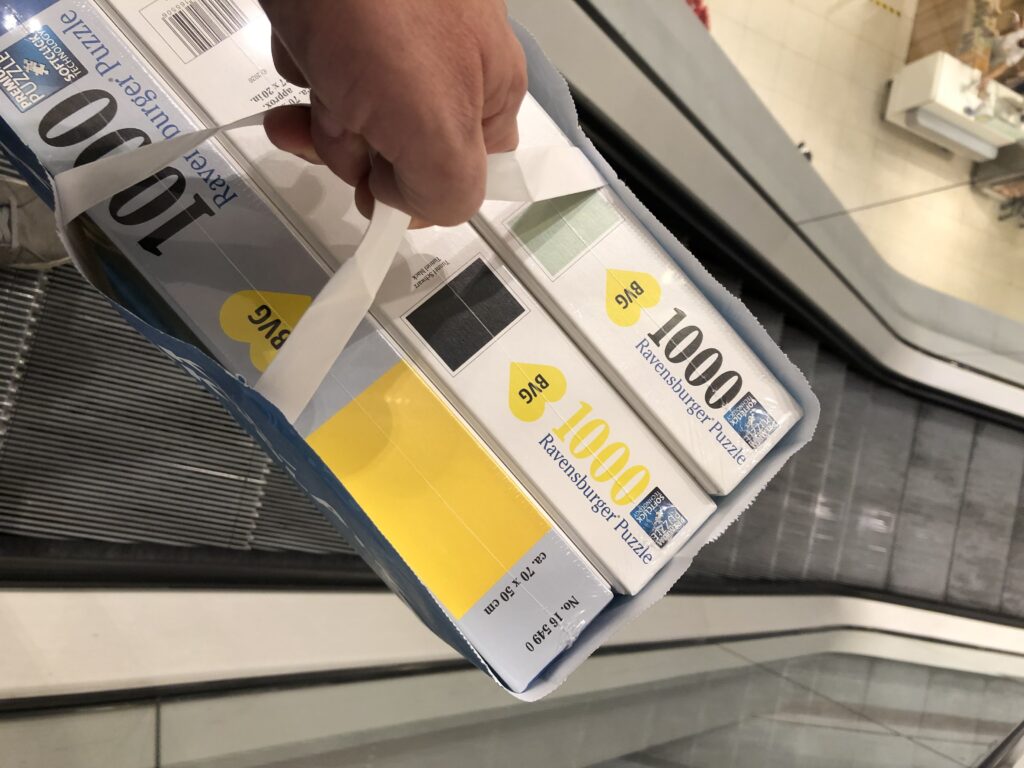 BVG Puzzle Paul Ripke - Eine Hand hält eine blaue Papiereinkaufstasche mit mehreren neuen Ravensburger Puzzleschachteln mit der Aufschrift "1000" und "Premium Puzzle", positioniert über einer Rolltreppe, die die Rolltreppe und die Deckenbeleuchtung widerspiegelt.