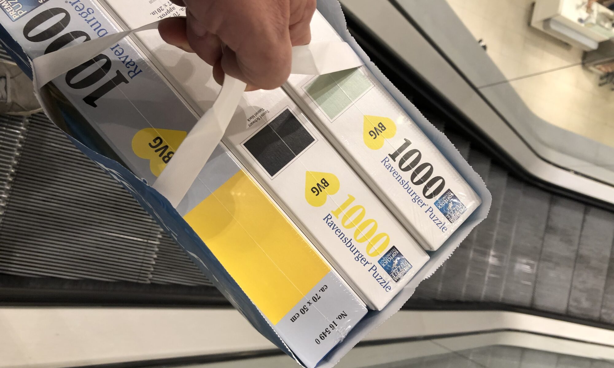 BVG Puzzle Paul Ripke - Eine Hand hält eine blaue Papiereinkaufstasche mit mehreren neuen Ravensburger Puzzleschachteln mit der Aufschrift "1000" und "Premium Puzzle", positioniert über einer Rolltreppe, die die Rolltreppe und die Deckenbeleuchtung widerspiegelt.