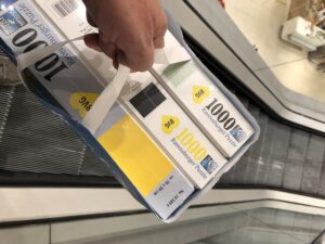 BVG Puzzle Paul Ripke - Eine Hand hält eine blaue Papiereinkaufstasche mit mehreren neuen Ravensburger Puzzleschachteln mit der Aufschrift "1000" und "Premium Puzzle", positioniert über einer Rolltreppe, die die Rolltreppe und die Deckenbeleuchtung widerspiegelt.
