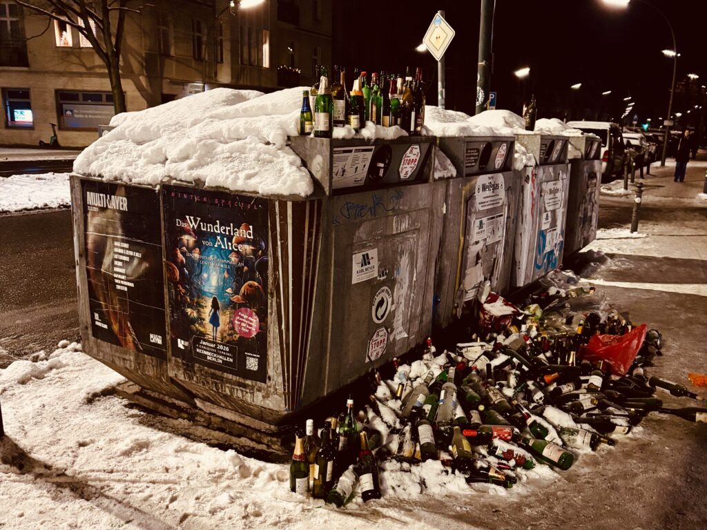 Flaschen, Fragen und Antworten - Eine nächtliche Straßenszene mit schneebedeckten Metallrecyclingcontainern, die mit weggeworfenen Glasflaschen überlaufen und mit Schnee bedeckt sind, beleuchtet von Straßenlaternen.