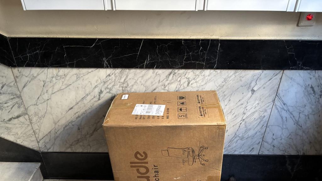 Spass mit DPD #4678 - Ein Pappkarton mit der Aufschrift "Holludle Schreibtischstuhl" und einer schematischen Darstellung eines Schreibtischstuhls steht auf einem Marmorboden vor einem kontrastierenden schwarzen Marmorakzent.