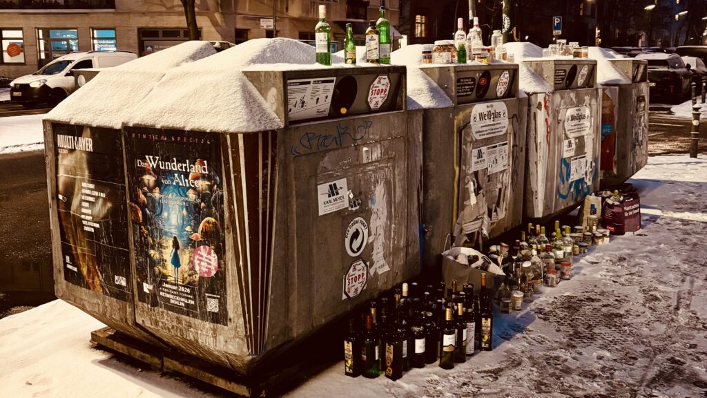 Wochenrueckblick kw 05 2026 su.day - Eine Reihe schneebedeckter Recyclingbehälter mit angebrachten Plakaten auf der Außenseite, die mit leeren Glasflaschen überfüllt sind, in einer verschneiten städtischen Umgebung, mit ein paar Autos, die im Hintergrund auf der Straße geparkt sind.