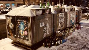 Wochenrueckblick kw 05 2026 su.day - Eine Reihe schneebedeckter Recyclingbehälter mit angebrachten Plakaten auf der Außenseite, die mit leeren Glasflaschen überfüllt sind, in einer verschneiten städtischen Umgebung, mit ein paar Autos, die im Hintergrund auf der Straße geparkt sind.