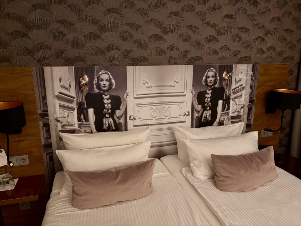 Wochenrueckblick KW 07 2026 - Ein Hotelbett mit zwei Einzelbetten, mit weißen Laken und Kissen sowie einem Akzentkissen aus Samt. Über dem Bett befindet sich ein Kopfteil mit einem Schwarz-Weiß-Bild einer Frau in einem Vintage-Kleid und einer verzierten Umgebung. Auf jeder Seite des Kopfteils befindet sich eine Lampe.