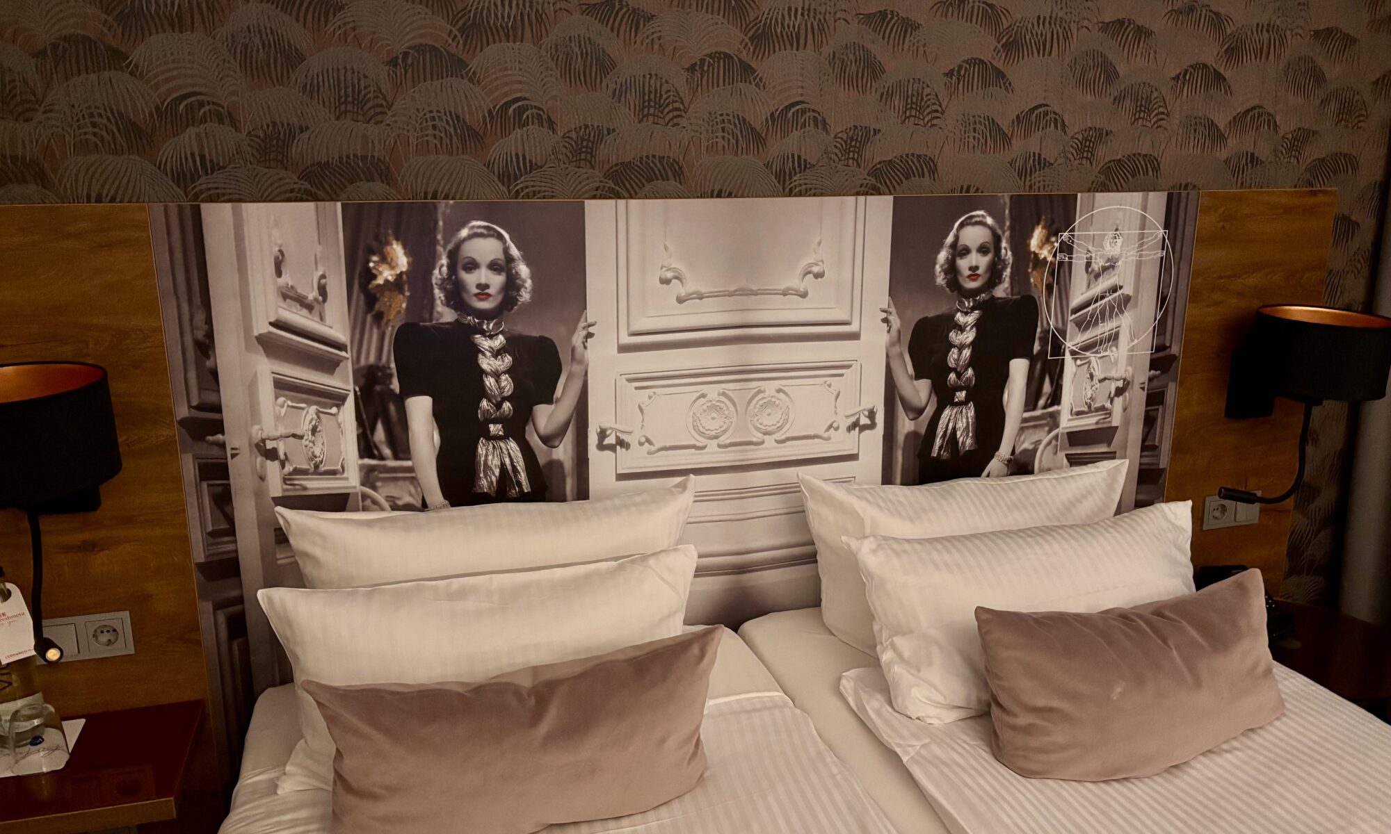 Wochenrueckblick KW 07 2026 - Ein Hotelbett mit zwei Einzelbetten, mit weißen Laken und Kissen sowie einem Akzentkissen aus Samt. Über dem Bett befindet sich ein Kopfteil mit einem Schwarz-Weiß-Bild einer Frau in einem Vintage-Kleid und einer verzierten Umgebung. Auf jeder Seite des Kopfteils befindet sich eine Lampe.