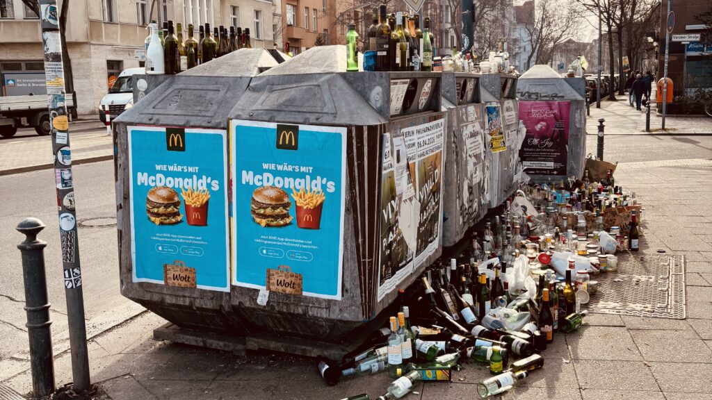 Wochenrueckblick kw 11 2026 slow motion - Große, überquellende öffentliche Recyclingbehälter in Berlin, Deutschland, bedeckt mit Werbung für McDonald's und Konzertplakaten, umgeben von weggeworfenen Flaschen und Müll auf dem Bürgersteig.