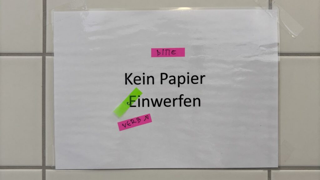 Wochenrueckblick kw 12 2026 yakle - A white sign with black text reads "Kein Papier Einwerfen" (No Paper to be thrown in). Pink sticky notes with "BITTE" (Please) and "VERB ↗" (Verb ↗) are attached to the sign. Ein weißes Schild mit schwarzer Aufschrift "Kein Papier Einwerfen". Daran befestigte rosa Haftnotizen mit "BITTE" und "VERB ↗".