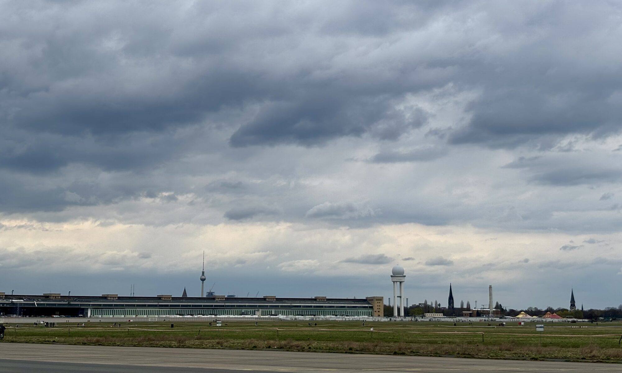 Wochenrueckblick kw 14 2026 der rentengaenger - Ein langes, niedriges Flughafenterminalgebäude erstreckt sich unter einem dramatischen, bewölkten Himmel über die Mitte des Bildes. Rechts vom Gebäude steht ein hoher, weißer Radarturm, und links ist in der Ferne der Berliner Fernsehturm sichtbar. Im Vordergrund sind grasbewachsene Felder zu sehen.