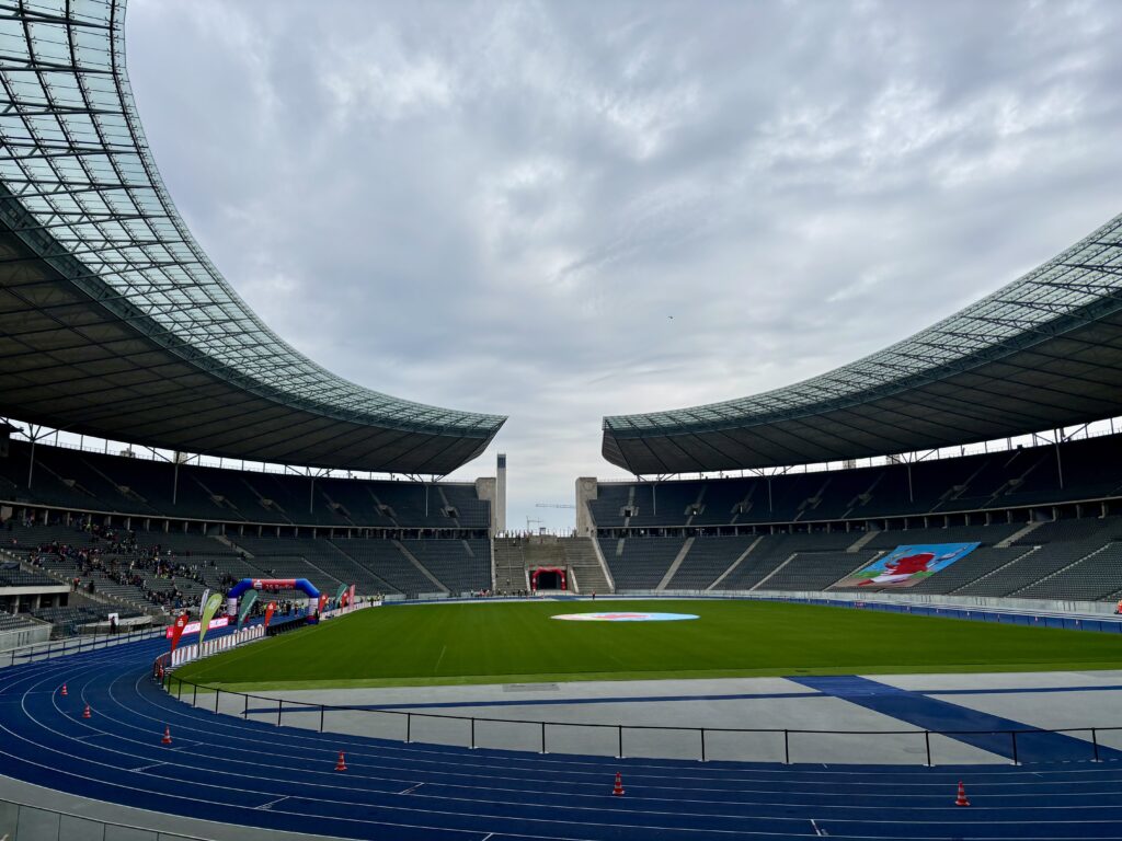 Wochenrueckblick kw 16 2026 arzttermin - olympiastadion - Eine weite Aufnahme eines großen modernen Stadions mit geschwungenen Dächern und gestuften Tribünen. Eine blaue Laufbahn umgibt einen grünen Rasenplatz. Bögen und Banner sind auf dem Spielfeld aufgebaut.