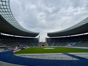 Wochenrueckblick kw 16 2026 arzttermin - olympiastadion - Eine weite Aufnahme eines großen modernen Stadions mit geschwungenen Dächern und gestuften Tribünen. Eine blaue Laufbahn umgibt einen grünen Rasenplatz. Bögen und Banner sind auf dem Spielfeld aufgebaut.