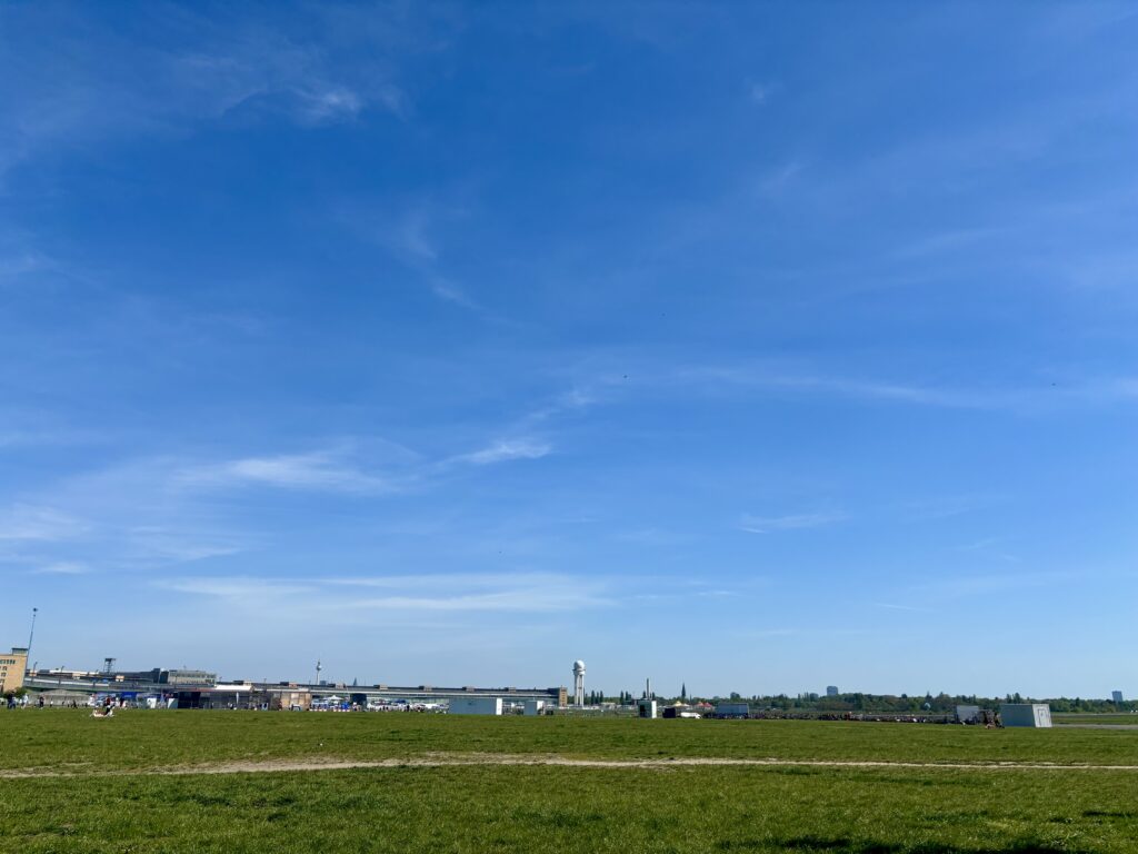Wochenrueckblick kw 18 2026 short tempelhofer feld - Ausgedehntes grünes Feld im Vordergrund unter einem strahlend blauen Himmel mit vereinzelten weißen Wolken. Im Mittelgrund erstreckt sich ein langer, niedrig-gebauter Gebäudekomplex horizontal, mit einem markanten runden Turm auf der linken Seite. Weitere Strukturen, darunter ein zylindrischer Turm und einige kleinere Gebäude, sind am Horizont zu sehen.