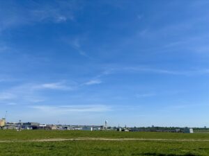 Wochenrueckblick kw 18 2026 short tempelhofer feld - Ausgedehntes grünes Feld im Vordergrund unter einem strahlend blauen Himmel mit vereinzelten weißen Wolken. Im Mittelgrund erstreckt sich ein langer, niedrig-gebauter Gebäudekomplex horizontal, mit einem markanten runden Turm auf der linken Seite. Weitere Strukturen, darunter ein zylindrischer Turm und einige kleinere Gebäude, sind am Horizont zu sehen.