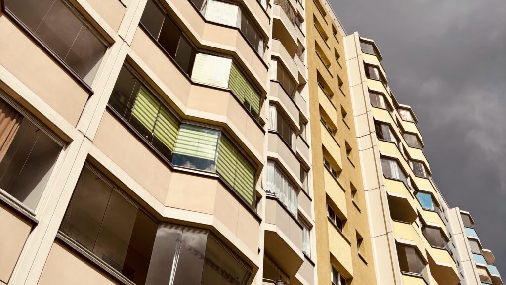 Wochenrueckblick kw 19 2026 yape plattenbau ost - Nahaufnahme eines hohen Wohngebäudes mit beigen und gelben Fassaden, das viele Fenster und Balkone vor einem dramatischen, dunklen Wolkenhimmel zeigt.