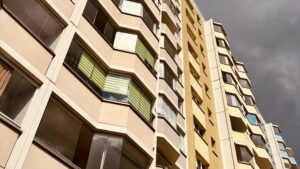 Wochenrueckblick kw 19 2026 yape plattenbau ost - Nahaufnahme eines hohen Wohngebäudes mit beigen und gelben Fassaden, das viele Fenster und Balkone vor einem dramatischen, dunklen Wolkenhimmel zeigt.