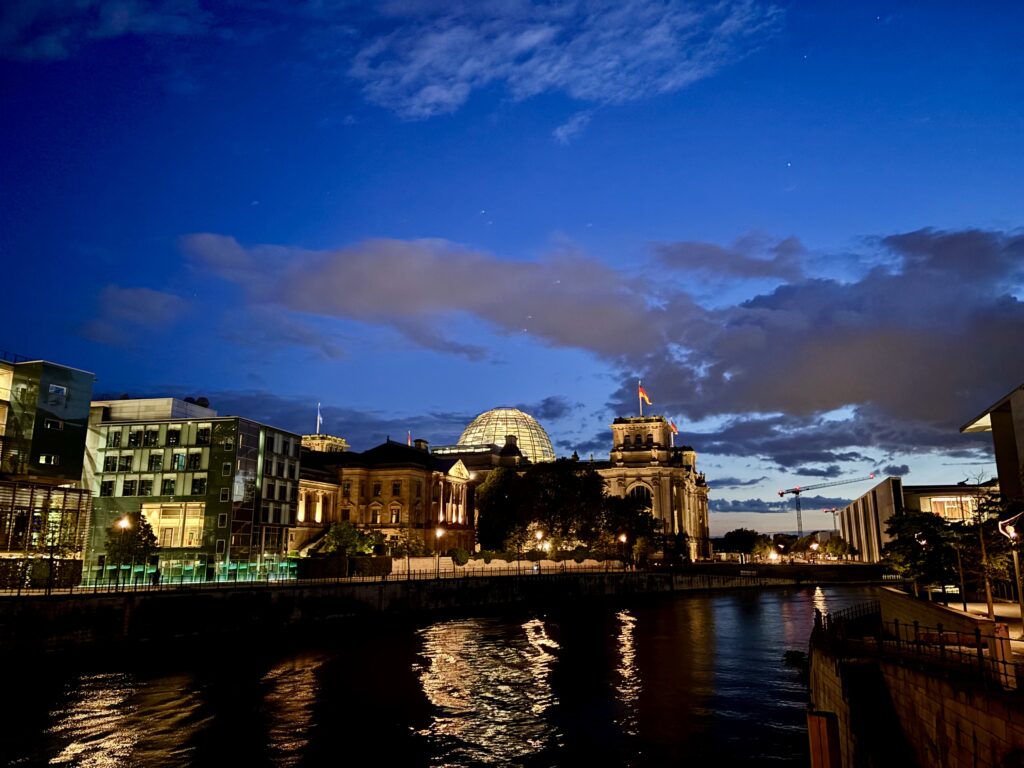 Wochenrueckblick kw 20 2026 krimi inside wie die touries - Reichstagsgebäude mit seiner Glaskuppel und Fahne in der Dämmerung, beleuchtet von Lichtern, die sich im Fluss Spree spiegeln, mit modernen Glasgebäuden und einem Baukran im Hintergrund unter einem dramatischen Himmel.