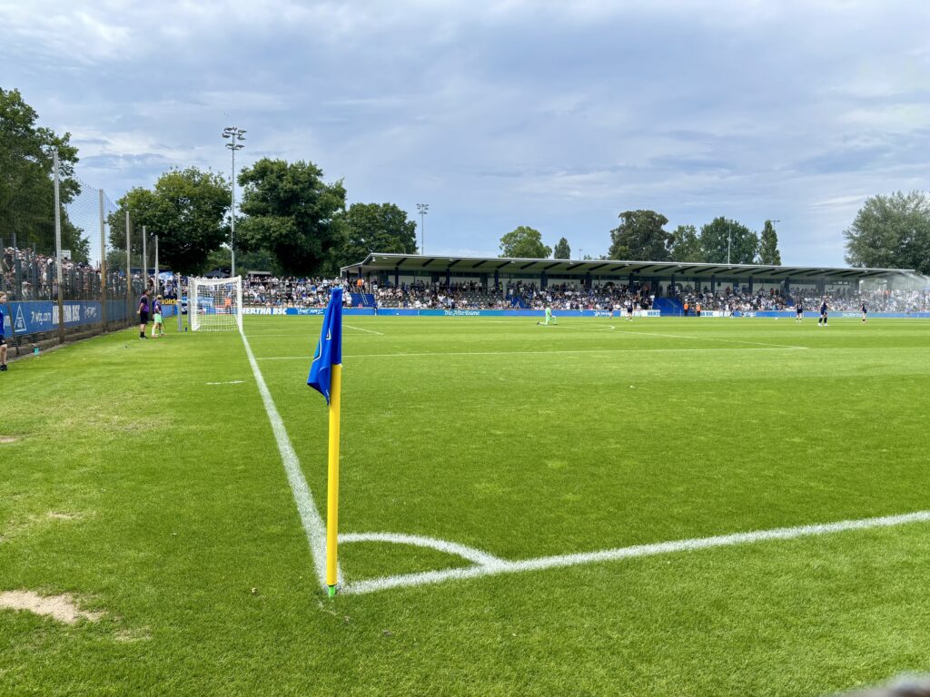 Wochenrueckblick kw 25 2026 hahohe - Eine blaue Eckfahne steht auf einem grünen Fußballfeld vor einem mit Zuschauern gefüllten Stadion unter einem bewölkten Himmel.