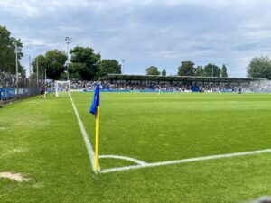 Wochenrueckblick kw 25 2026 hahohe - Eine blaue Eckfahne steht auf einem grünen Fußballfeld vor einem mit Zuschauern gefüllten Stadion unter einem bewölkten Himmel.