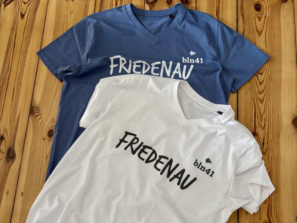 Friedenau again - Zwei V-Ausschnitt-T-Shirts, eines blau und eines weiß, jeweils bedruckt mit "FRIEDENAU" und einem kleinen Logo, auf dem "bln41" steht, auf einem Holzhintergrund.
