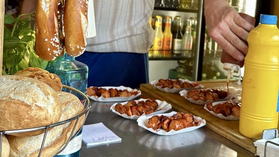 Wochenrueckblick kw 27 2026 tdc tour de currywurst - Gegrillte Würstchen in Papierbooten, auf die eine Hand Soße träufelt, daneben ein Brotkorb, aufgehängte Brezeln und eine große Senfflasche an einem Essensstand.