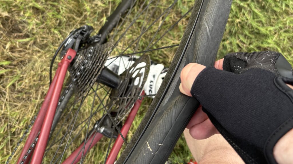 Nahaufnahme einer Hand, die einen schwarzen Fingerling trägt und einen Fahrradreifen drückt, wobei das Fahrrad seitlich auf grasigem Boden liegt und das Schaltwerk, die Kassette und die Speichen zu sehen sind.