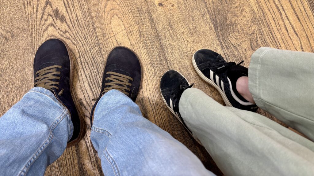 Nahaufnahme von zwei Paar Füßen, die auf einem Holzboden lässige Schuhe tragen. Das linke Paar trägt dunkelblaue Schnürschuhe mit senfgelben Schnürsenkeln und hellblaue Jeans. Das rechte Paar trägt schwarze Adidas-Sneaker mit weißen Streifen und hellgrüne Hosen.