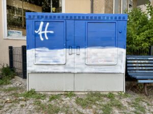 A blue and white utility box with graffiti art resembling clouds and a white "H" spray-painted on the top half, situated on a cobblestone path beside a bench and greenery. Eine blaue und weiße Verteilerkasten mit Graffiti-Kunst, die Wolken und ein weißes "H" auf der oberen Hälfte ähneln, auf einem Kopfsteinpflasterweg neben einer Bank und Grünpflanzen.