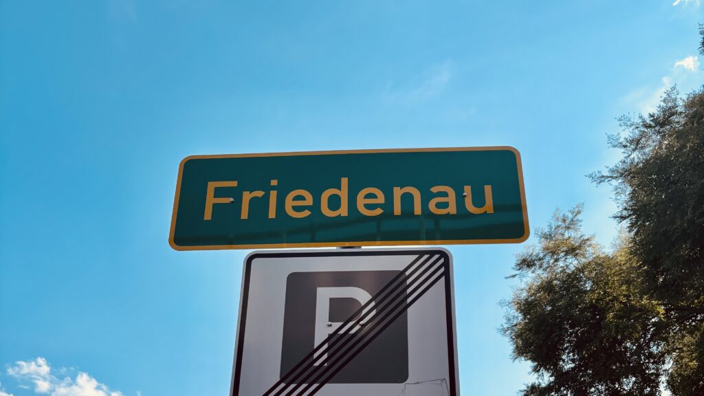 Green street sign with yellow text "Friedenau" and an end-of-parking sign below against a blue sky with trees on the right. Grünes Straßenschild mit gelbem Text "Friedenau" und einem Parkverbotsschild darunter vor einem blauen Himmel mit Bäumen rechts.