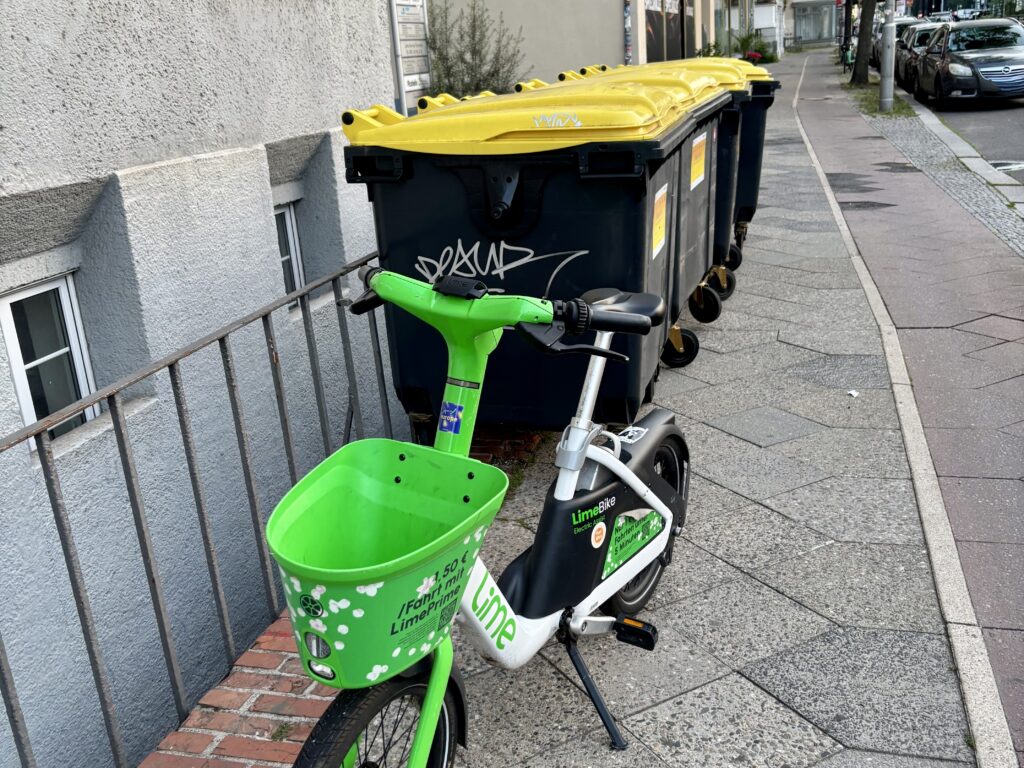 A bright green electric scooter with a basket stands on a sidewalk next to a row of large, dark gray trash bins with yellow lids, some marked with graffiti. Ein knallgrünes Elektroscooter mit Korb steht auf einem Bürgersteig neben einer Reihe von großen, dunkelgrauen Mülltonnen mit gelben Deckeln, einige mit Graffiti versehen.