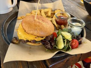 A juicy cheeseburger topped with a wooden skewer, served with golden french fries, a small side salad of mixed greens, tomatoes, and cucumbers, a small jar of ketchup, and a small jar of mayonnaise on a brown paper-lined tray. Ein saftiger Cheeseburger, garniert mit einem Holzspieß, serviert mit goldenen Pommes Frites, einem kleinen Beilagensalat aus gemischtem Grün, Tomaten und Gurken, einem kleinen Glas Ketchup und einem kleinen Glas Mayonnaise auf einem Tablett mit braunem Papier ausgelegt.